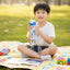 Child sitting on a colorful blanket in a park holding a water bottle.